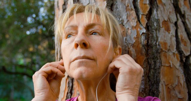 Senior woman adjusting her earphones while resting against a tree in nature. Use for topics on active aging, outdoor relaxation, fitness for seniors, and peaceful moments in nature.