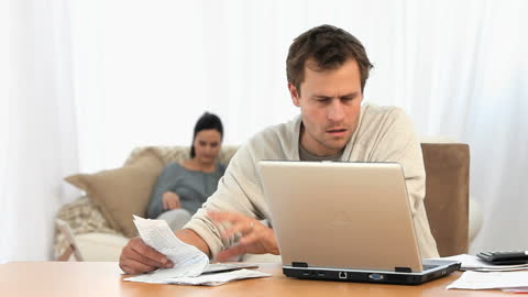 Concerned Man Calculating Bills with Family in Background at Home