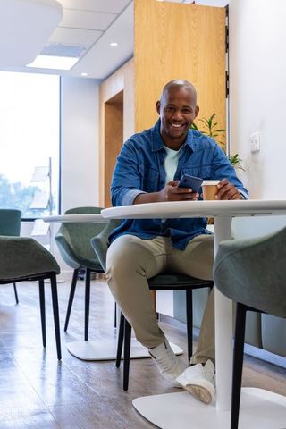 Smiling african man relaxing in modern coworking lounge with coffee and smartphone