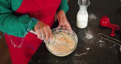 Whisking batter in red apron and green sweater on dark countertop with milk and flour