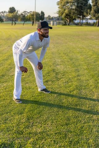 Crouching cricketer in white uniform holding red ball on sunlit grass field