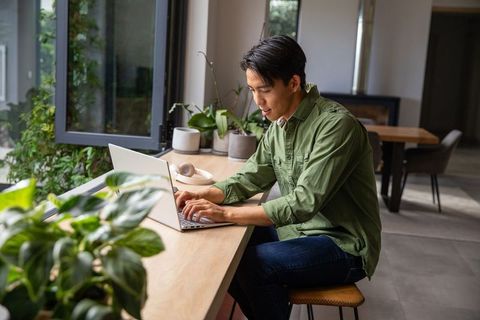 Young asian man typing on laptop at sunlit home counter surrounded by plants and headphones