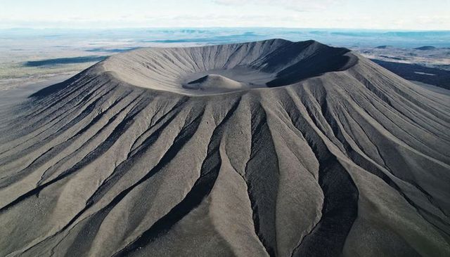 Aerial view showing circular cinder cone with radial gullies on arid plain under pale sky