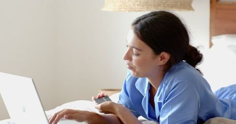 Woman in Blue Pajamas Using Laptop and Phone in Cozy Bedroom Setup