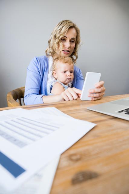 Mother and baby sitting at table and using mobile phone at home