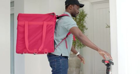 African american courier carrying red insulated backpack reaching scooter at home entrance