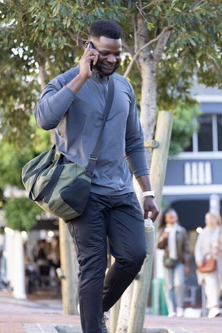 Smiling african man walking tree-lined sidewalk talking on phone carrying gym duffel and bottle