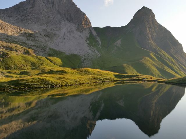 Beautiful, calm scene of majestic mountains reflected in crystal clear lake. Green rolling hills gently slope to water's edge, touched by warm sunlight. Perfect for travel blogs, nature websites, prints for decor, meditation material, and posters emphasizing peaceful, serene environments.
