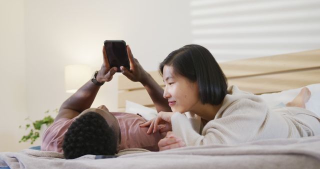 Couple lying in bed, sharing a relaxing moment while using their smartphone together. Great for illustrating themes of modern technology in everyday life, interracial relationships, comfort at home, and bedroom relaxation.
