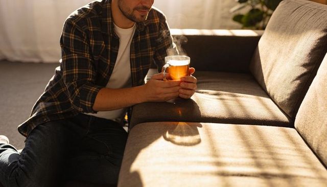 Kneeling man holding steaming glass mug by sofa in sunlit cozy living room wearing flannel