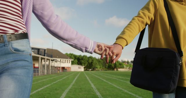 A couple is holding hands while walking on a public field on a sunny day. Both individuals are dressed in casual outfits. This image can be used for websites or campaigns promoting romance, relationships, outdoor activities, or lifestyle themes. It can also be suitable for blog posts about love, companionship, or enjoying nature together.