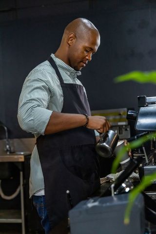 Barista pouring steamed milk into to-go cup at espresso machine in moody industrial cafe