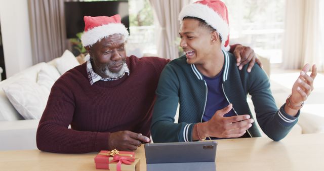 Father and son are wearing Christmas hats. They are sharing a joyful moment while using a tablet for a video call. A wrapped gift with a red ribbon is displayed on the table. This image conveys a festive atmosphere, ideal for promoting family-oriented Christmas celebrations, holiday greetings, or technology use during festive times.