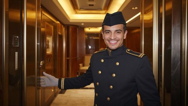 Smiling bellman welcoming guest at luxury hotel elevator with polished brass doors and gold trim