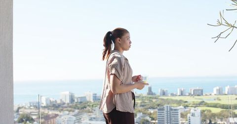 Woman standing on balcony holding cup overlooking coastal city skyline