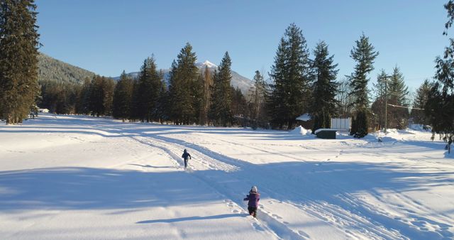 Two children walk through a snow-covered field surrounded by trees and cabins. The scene is set against a crisp blue sky with mountain views in the background. Ideal for use in illustrating winter activities, holiday postcards, seasonal greetings, or articles about outdoor fun and nature.