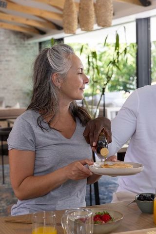 Mature woman enjoying pancake while partner pours syrup at sunlit rustic breakfast table