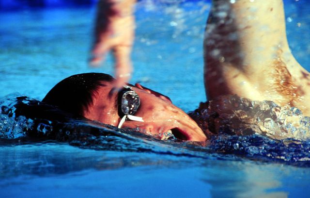 This image shows a professional swimmer competing in a race. He is moving through the water with strong strokes, wearing swim goggles and displaying determination. It is suitable for promoting swimming competitions, athletic events, sports training programs, swimwear advertisements, or fitness campaigns.