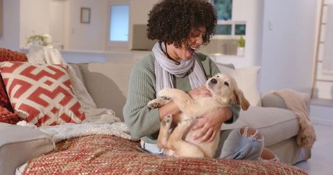 African american woman cradling golden retriever puppy on cozy living room sofa, smiling