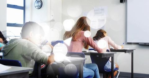 Teen students writing at desks in bright classroom with natural window light and notebooks
