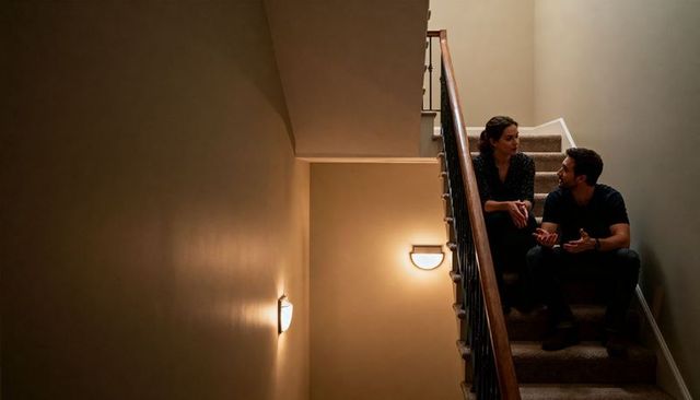 Couple talking on carpeted stair landing with warm wall sconce lighting, wooden banister