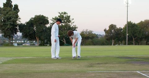 Two adult male cricket teammates inspecting pitch crease on grassy field at dusk