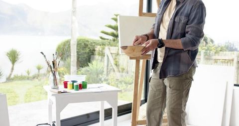 Indian artist mixing paint in sunlit studio by glass doors with easel and garden view