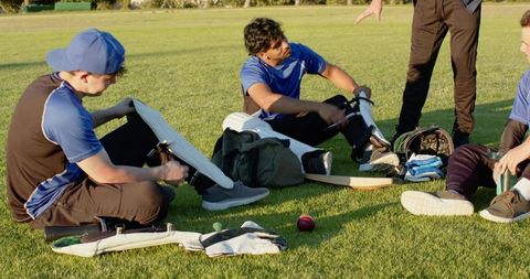 Diverse teammates adjusting cricket pads and inspecting gear on sunlit grassy field