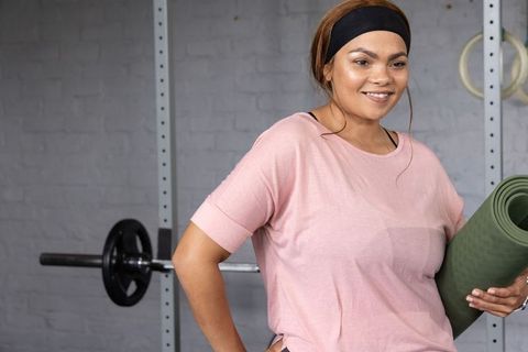 Smiling woman holding green yoga mat near squat rack and barbell in modern gym