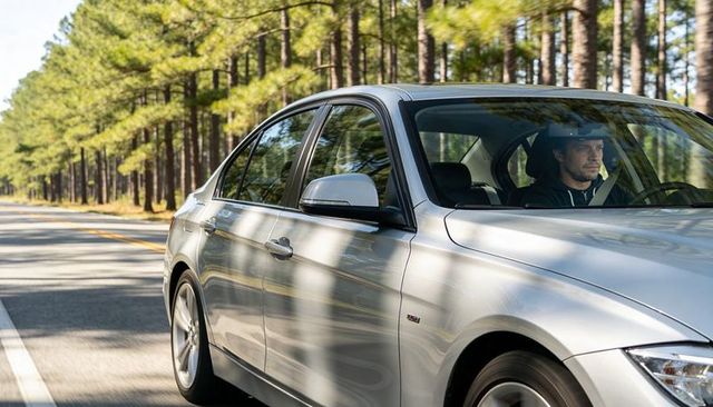 Silver sedan driving along sunlit pine forest road with focused male driver wearing hoodie