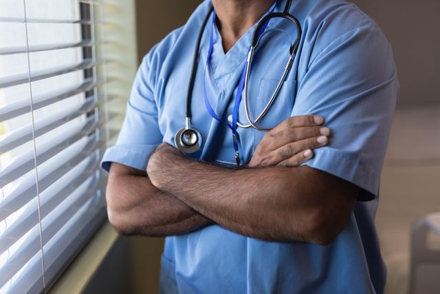 Mid section of biracial male doctor standing with arms crossed at hospital
