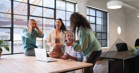 Laughing coworkers celebrating success around wooden table in modern office with open laptop