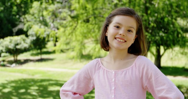 Young girl standing in a park on a sunny day, smiling and looking at camera. She is in a nature setting with vibrant green trees and grass in the background. Perfect for use in advertisements, promotional materials, or websites related to children's activities, outdoor fun, happiness, and leisure.