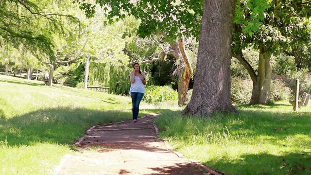 Young woman engaged in a phone conversation while walking through a lush green park. Surrounded by leafy trees and sunlight, she's enjoying leisure time and nature. Perfect for illustrating concepts of communication, relaxation, or outdoor activities in a natural setting.