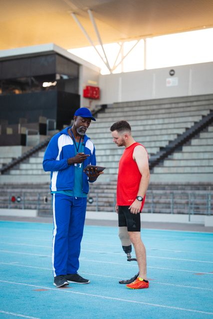 Coach and athlete standing on a track, discussing training plans while looking at a tablet. The athlete has a prosthetic leg, highlighting inclusivity and determination. Useful for themes related to sports coaching, professional training, fitness technology, and teamwork. Ideal for articles, advertisements, and promotional materials focusing on athletic training, sports mentorship, and adaptive sports.