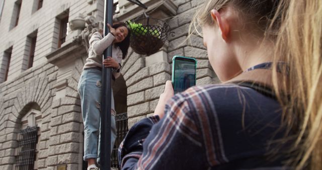 Teen girl captures candid photo of her friend climbing lamp post on a city street. Urban vibes and playful moment portray youthful energy and friendship. Perfect for content related to teen activities, friendship, urban exploration, and candid photography.