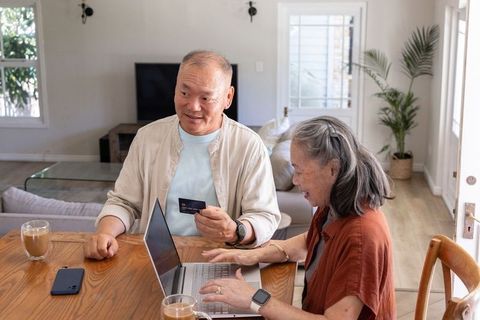 Senior asian couple making online purchase at home using laptop and credit card, smiling