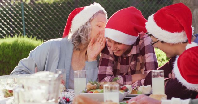 Grandmother whispering to her grandchildren while they share an outdoor Christmas meal. Everyone wears Santa hats and the table is filled with festive food and drinks. Ideal for holiday promotions, family-oriented advertisements, or content focusing on holiday traditions and family bonds.