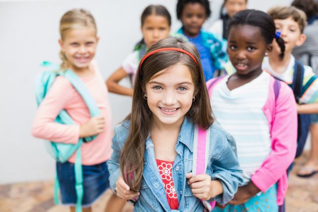 This image depicts a diverse group of cheerful school children standing outdoors with backpacks, smiling and ready for school. Ideal for use in educational materials, school advertising, websites promoting diversity and inclusion, and any context emphasizing childhood, friendship, or the beginning of a school year.