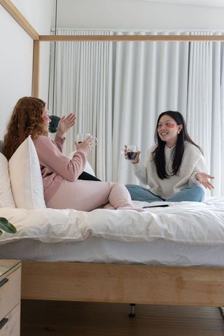 Mixed-ethnicity friends chatting on canopy bed sipping wine and coffee in cozy minimalist bedroom