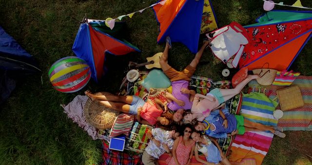 Group of friends resting on a picnic blanket, surrounded by colorful tents and cheerful summer decorations, enjoying a casual and joyful moment together. Suitable for promoting camping gear, outdoor activities, summer festivals, or picnic-related products. Can be used in advertisements or editorial content depicting friendship, leisure activities, and outdoor adventures.