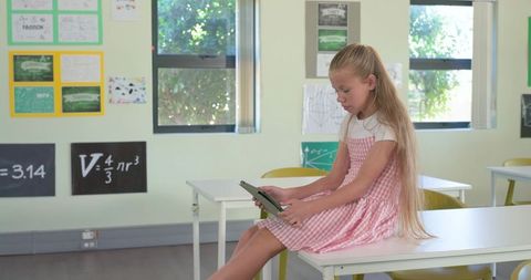 Preteen girl sitting on classroom desk using tablet wearing pink dress focusing on digital learning