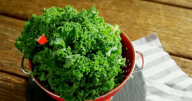 A red colander filled with fresh, green kale sits on a wooden table, with copy space. Kale is a nutritious leafy green vegetable often used in salads and healthy cooking.