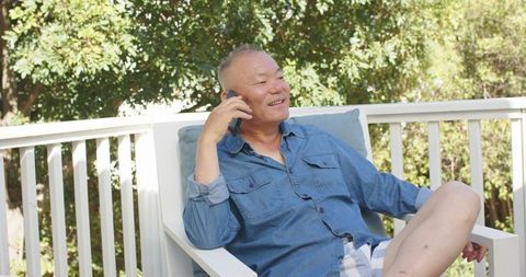 Senior asian man relaxing on sunlit deck talking on smartphone wearing denim shirt