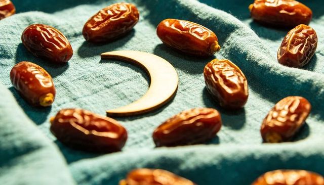 Ramadan crescent wooden ornament with glossy dates on teal linen for festive still life