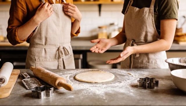 Bakers kneading dough together wearing linen aprons in cozy kitchen with rolling pin