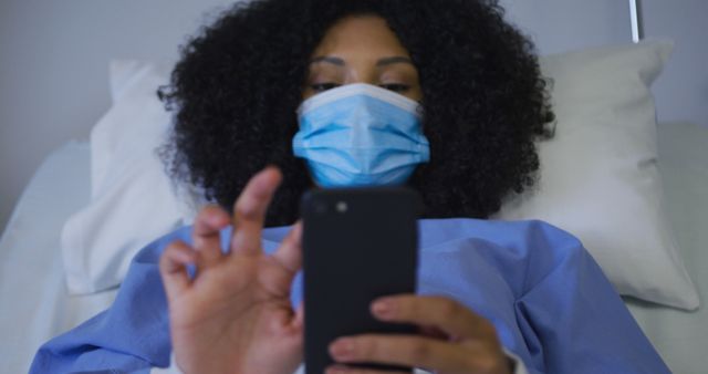African american female patient wearing face mask lying on hospital bed using smartphone. medicine, health and healthcare services.