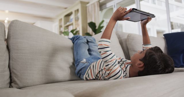 A young boy using a tablet while lying on a couch at home. The relaxed, casual setting makes it ideal for illustrating home lifestyle, children's activities, tech-savvy youth, or digital leisure. Suitable for blogs, articles, educational materials, and advertisements focused on technology, children, and modern family life.