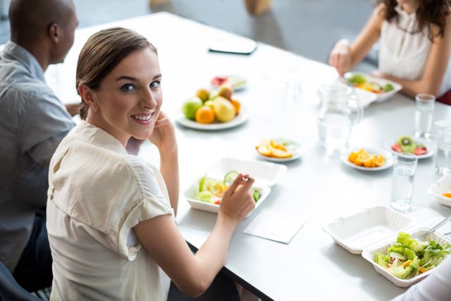 Businesswoman smiling while enjoying a healthy lunch in an office environment. Ideal for use in articles about workplace wellness, corporate culture, healthy eating habits, and team building activities.