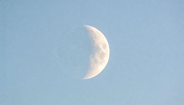 Daytime crescent moon revealing crater detail against pale blue sky minimalist celestial